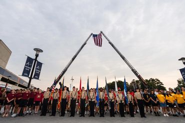 Group of scouts and participants under an American flag held by two fire truck ladders.