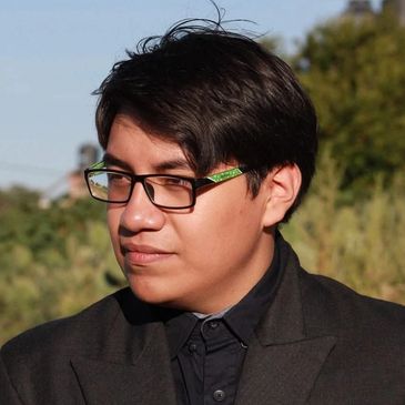 Young man wearing glasses and a black jacket looking to the side outdoors.