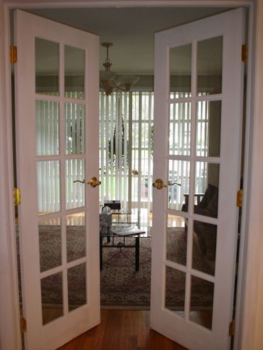 Open French doors reveal a cozy living room with a glass coffee table and vertical blinds.