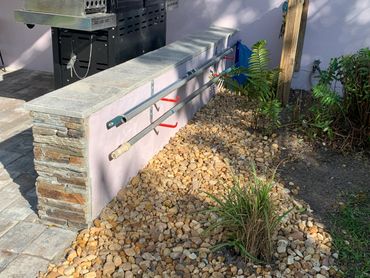 Outdoor area with stone wall, gravel bed, plants, and two metal poles mounted on wall.