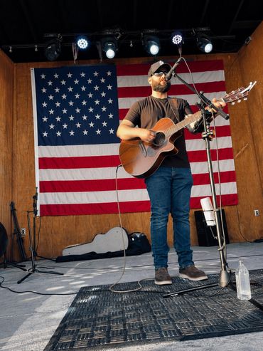 Man playing guitar and singing on stage with American flag backdrop.