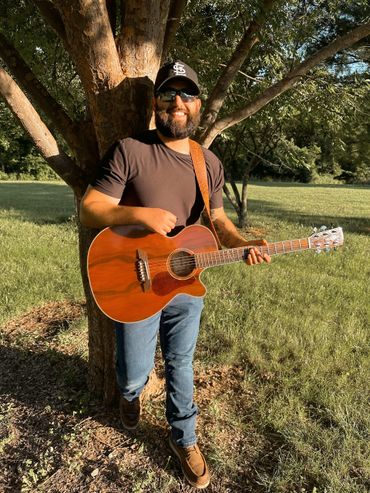 Man playing guitar outdoors, standing by a tree with sunglasses and a cap.