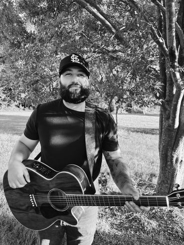 Man playing acoustic guitar outdoors under a tree.