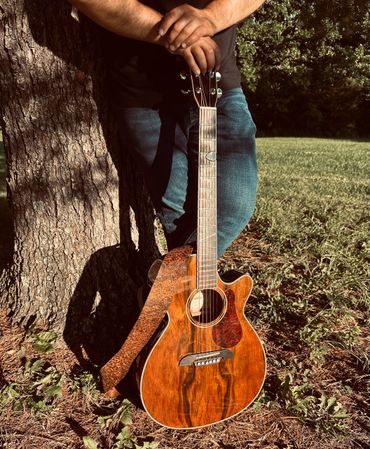 Person leaning on a tree holding an acoustic guitar outdoors.
