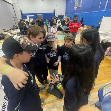 Kids in a gymnasium huddle together, wearing matching shirts.