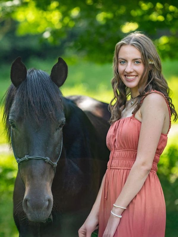 Young woman in a coral dress stands beside a black horse in a sunlit green field.