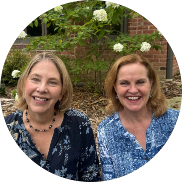 Two women smiling outdoors with flowers and greenery behind them.