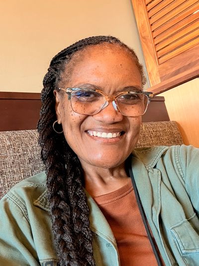 A smiling woman with braided hair and glasses sitting indoors.