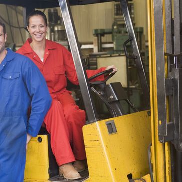 Two industrial workers smiling in a factory, one operating a forklift.