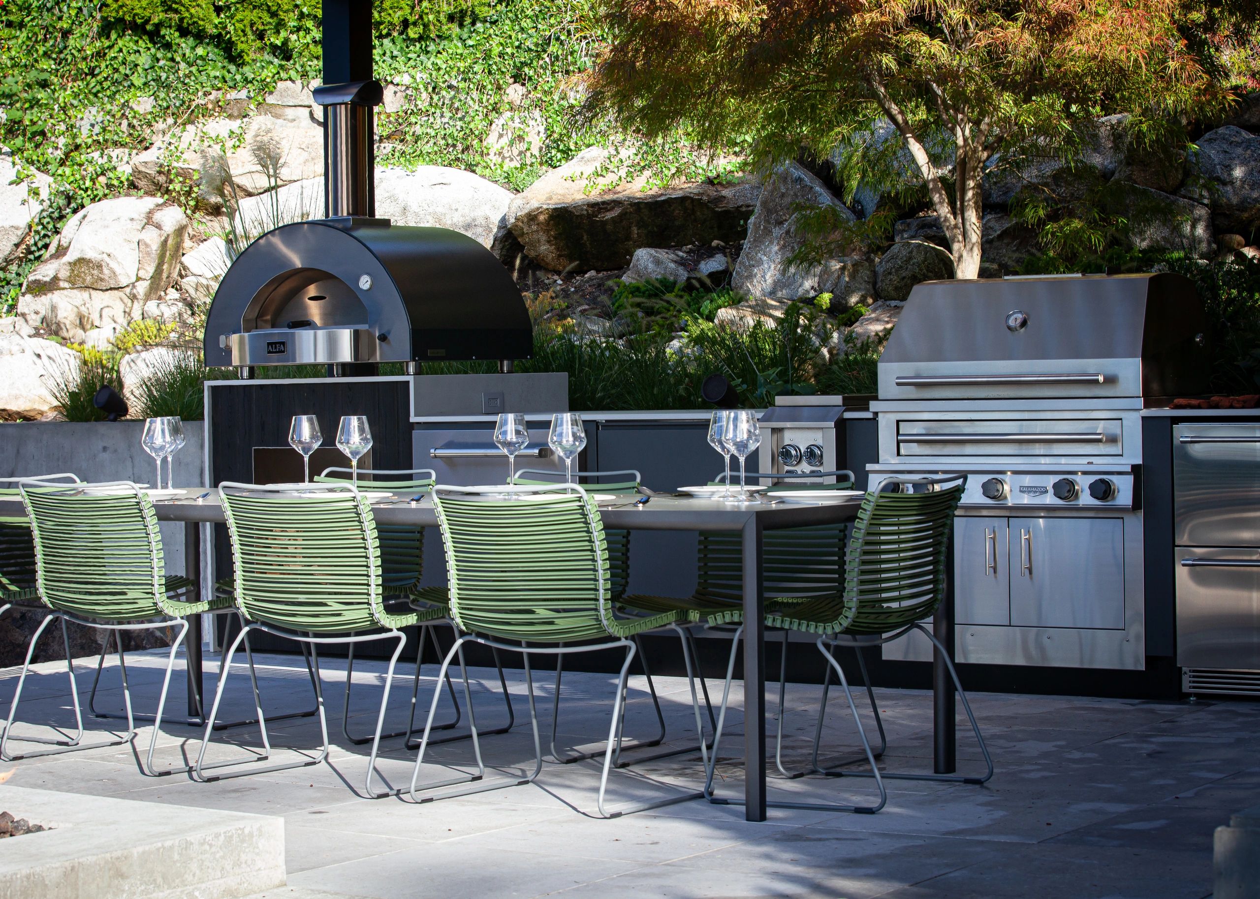 Outdoor dining area with green chairs, a wood-fired oven, and a stainless steel grill.