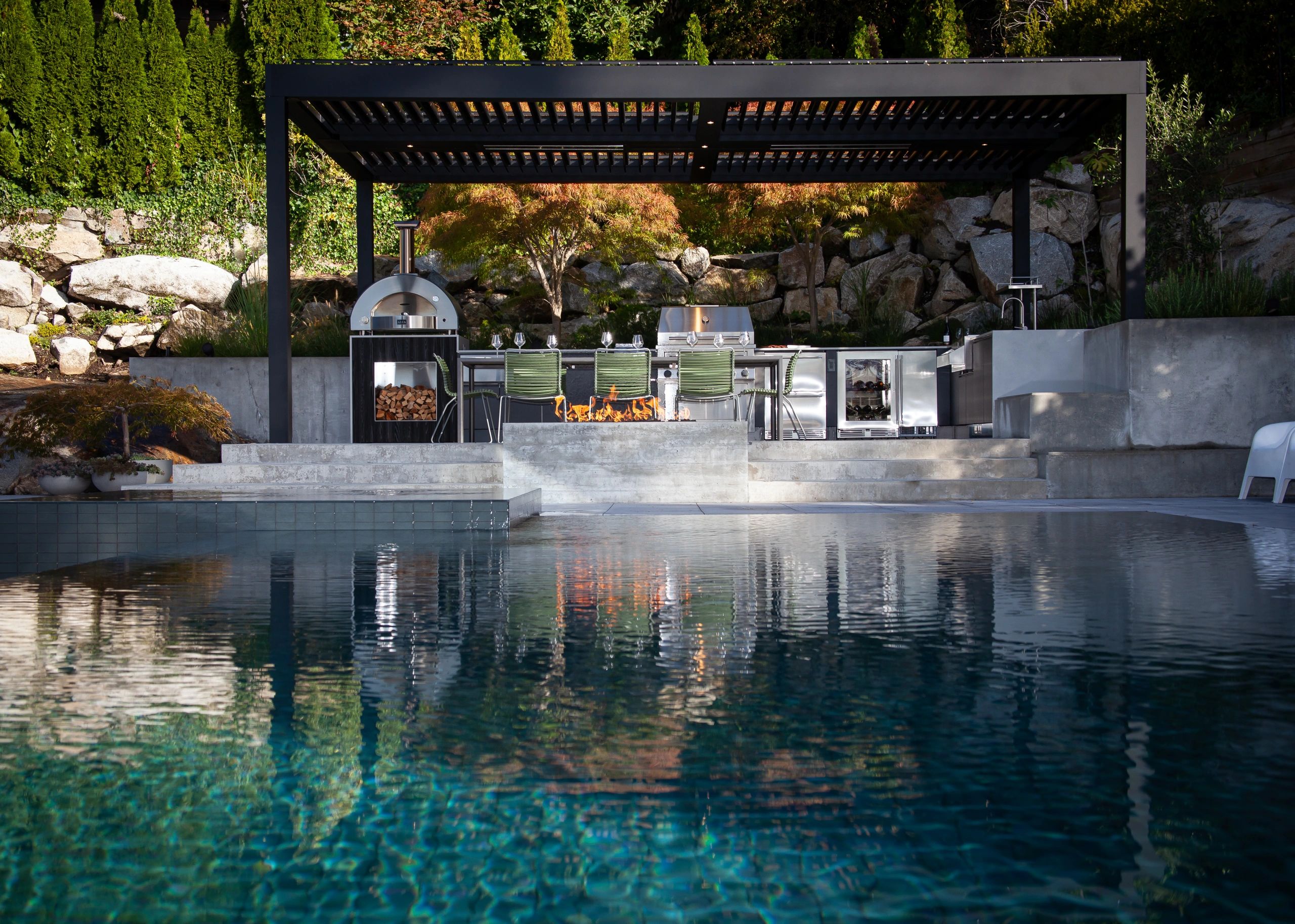 Modern outdoor kitchen with pergola, fireplace, and pool reflection.