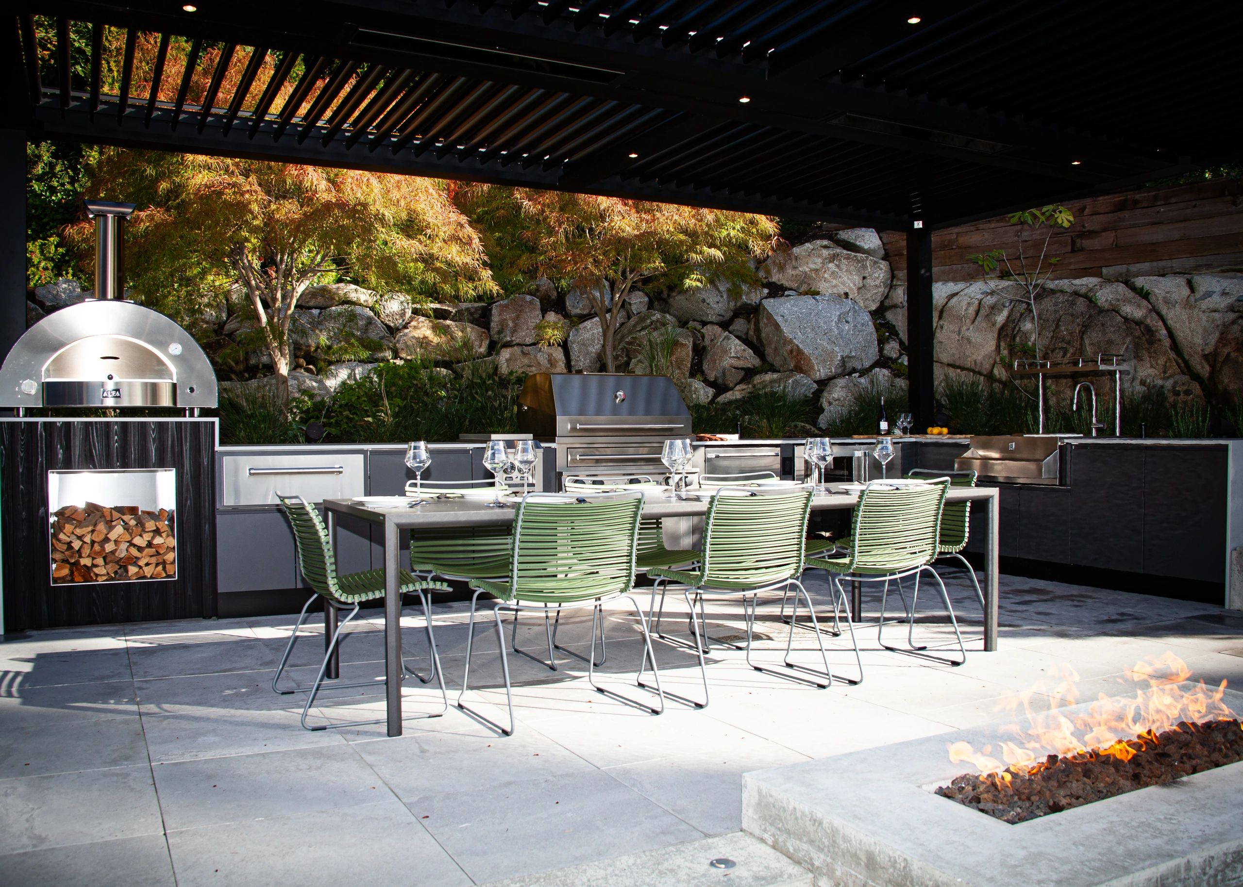 Modern outdoor kitchen and dining area with fire pit and greenery.