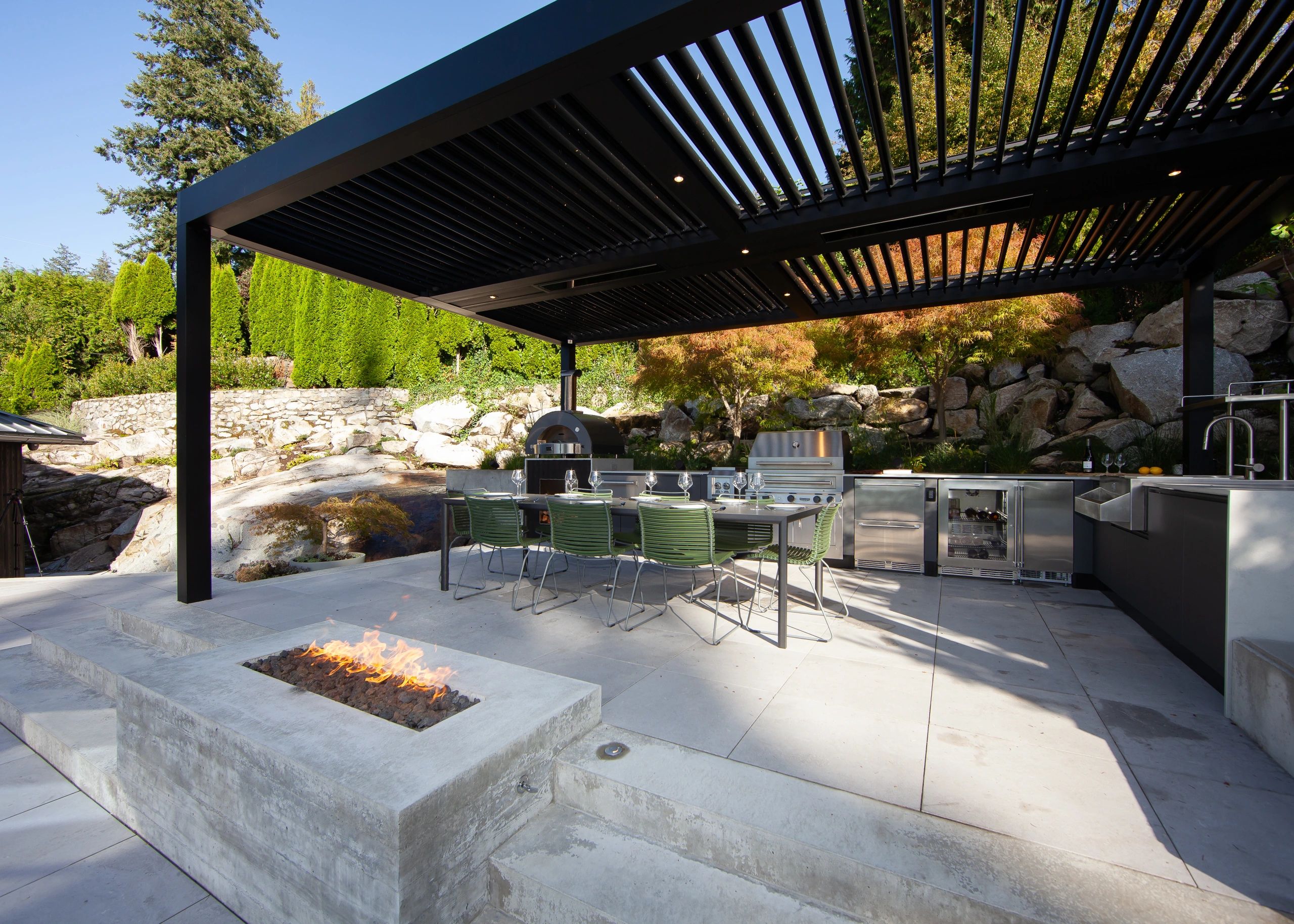 Modern outdoor kitchen with pergola, dining area, and fire pit in a natural setting.
