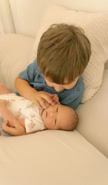 Older sibling gently touching newborn's face on a cozy couch.