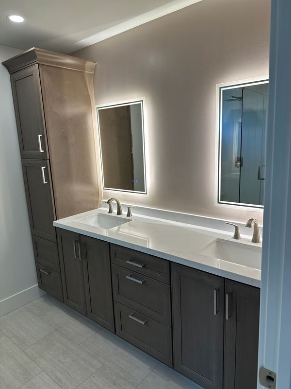 Brown cabinetry in master bathroom. Light up mirrors at double sink vanity. By Justice Renovations