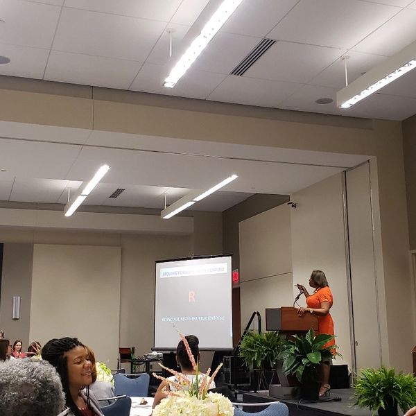 Speaker in orange presenting at a conference with attendees seated.