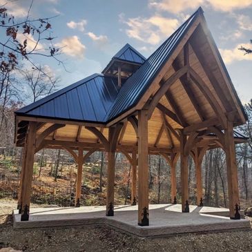 Wooden pavilion with a metal roof in a forest setting during sunset.