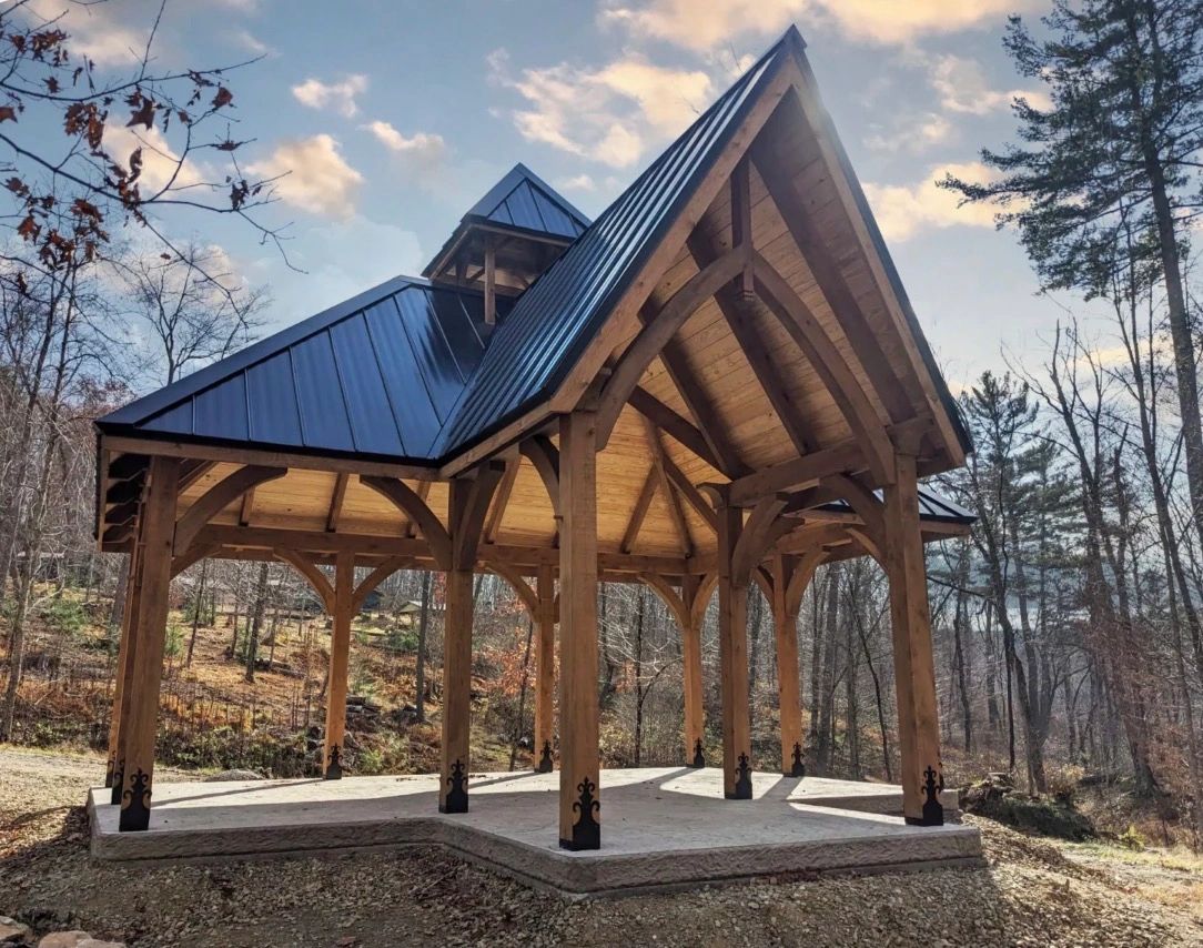A wooden pavilion with a black metal roof in a forest clearing.
