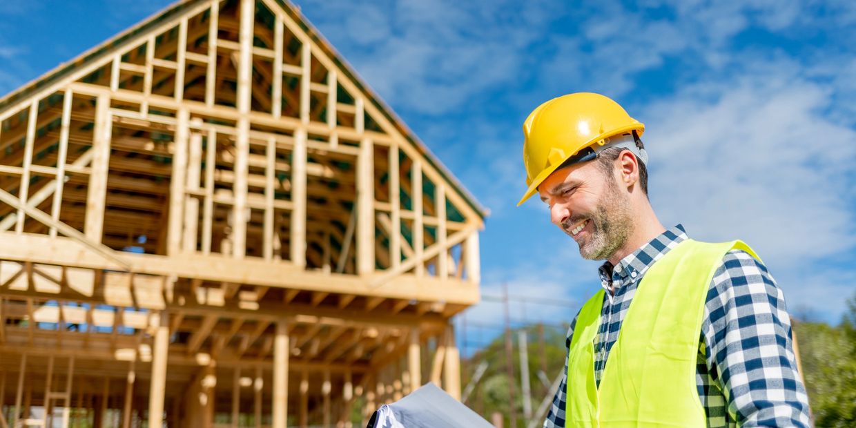 Construction worker reviewing plans at a wooden house frame under blue sky.