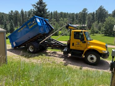 Yellow truck unloading a blue dumpster in a forested area.