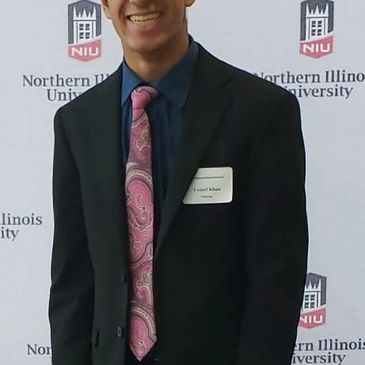 Yousuf Khan posing in a suit and tie in front of a NIU banner