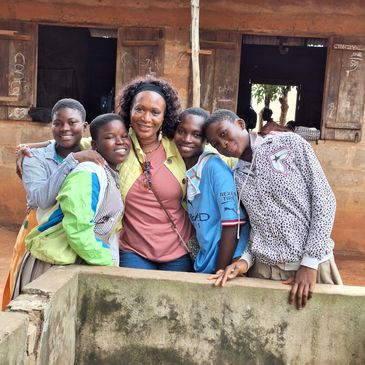 A group of smiling children and a woman posing together outside a rustic building.