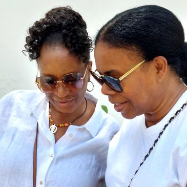 Two women wearing sunglasses, smiling and looking down together.