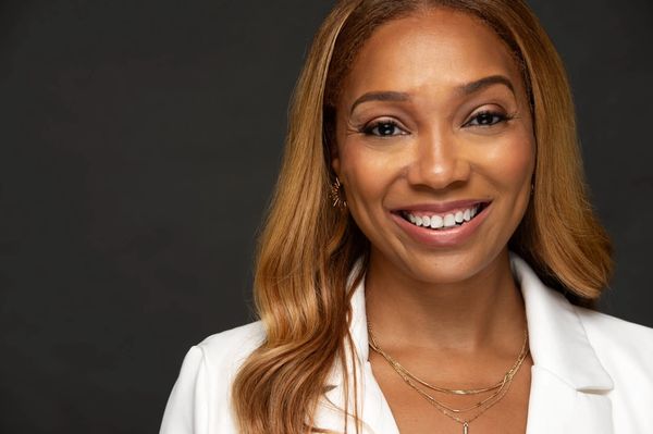 Smiling woman with long hair wearing a white blazer and gold necklaces.