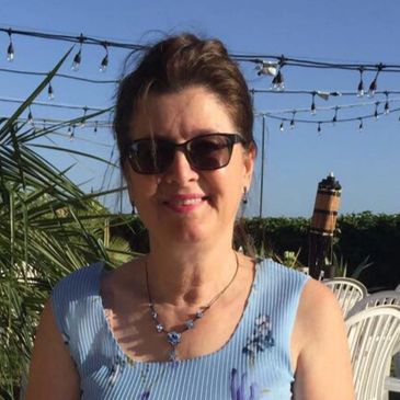 Smiling woman in sunglasses outdoors with string lights and greenery behind her.