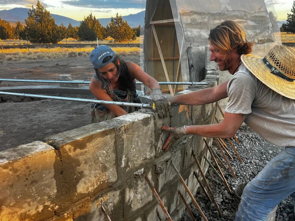 Two people building a concrete block wall outdoors during sunset.