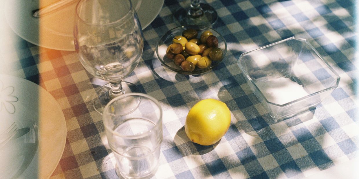 A lemon and bowl of olives on a blue checkered tablecloth with empty plates. By Roberta Sant'Anna