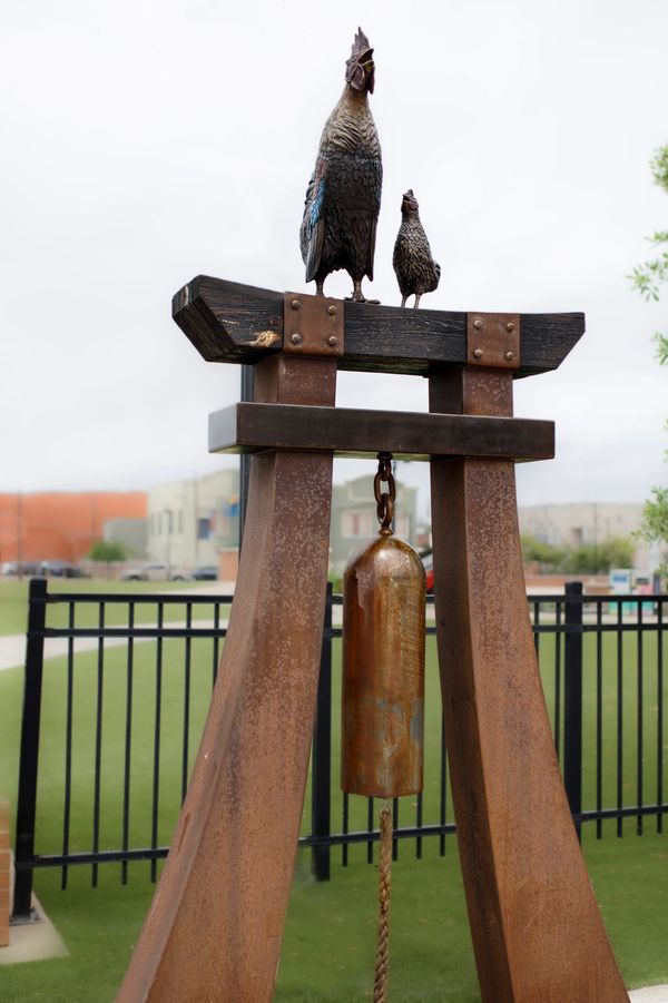 Metal rooster sculptures atop a rusted bell structure in a fenced outdoor area.
