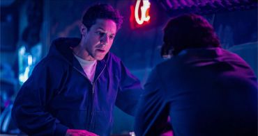 Two men converse in a dimly lit room with blue and red lighting.