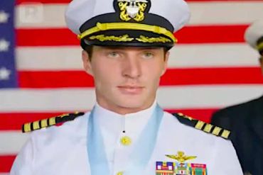 Naval officer in uniform with medal and flag backdrop.