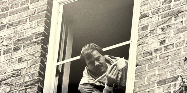 Early PACER photo: a painter leaning out of an upstairs window of a brick building, brush in hand