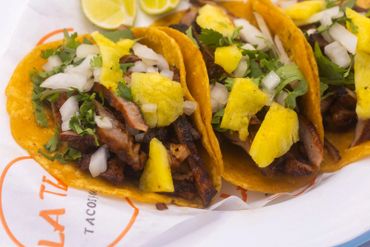 Three tacos filled with meat, pineapple, onions, and cilantro on a branded plate.