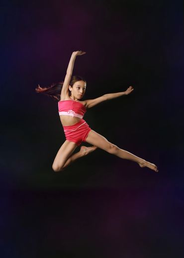 Young girl in pink outfit performing a mid-air dance leap against a dark background.