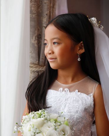Young girl in a white dress holding a bouquet, gazing out a window.
