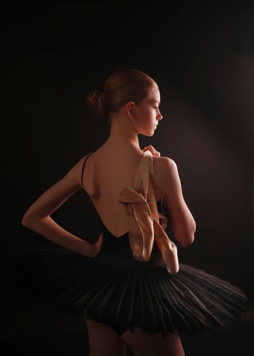 Ballerina in black tutu holding pointe shoes against a dark background.