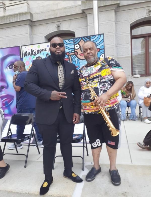 Two men posing outside, one in a suit and the other holding a saxophone.