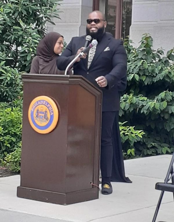 Man in sunglasses speaking at a Philadelphia podium outdoors.