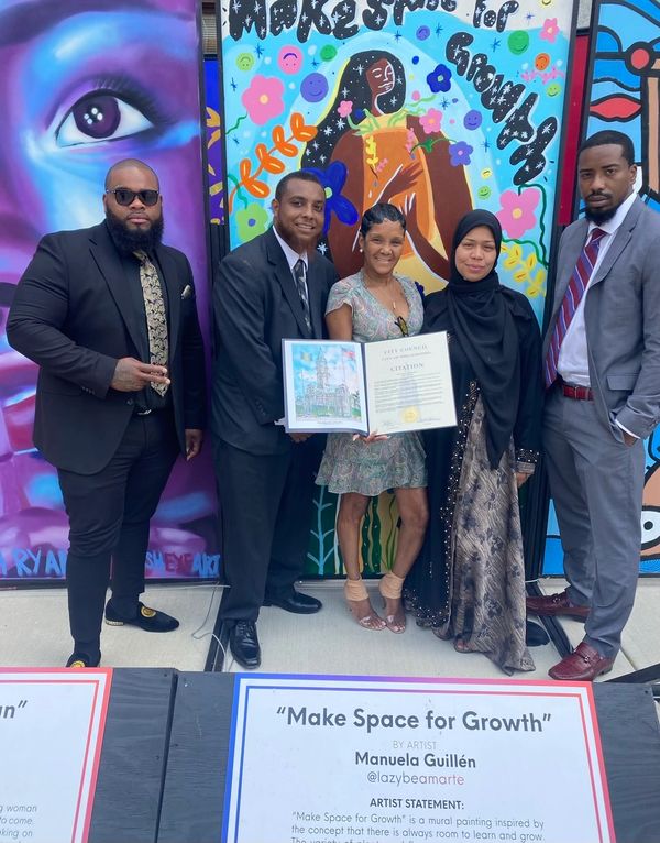 Group of five people posing with a city council citation in front of a colorful mural.