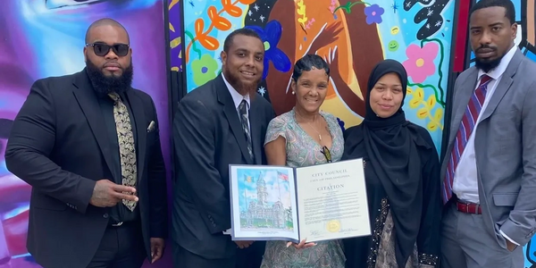 Five people in formal attire holding a city council citation in front of a colorful mural.