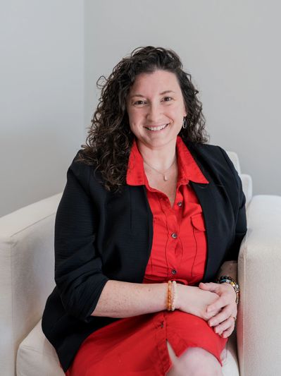 Smiling woman in a red dress and black blazer sitting on a white chair.