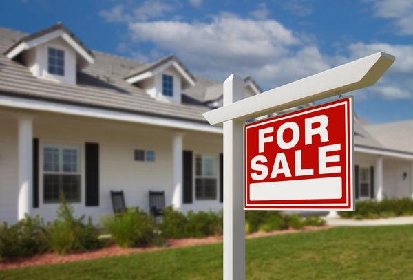 A "FOR SALE" sign with a red background stands in front of a modern house.