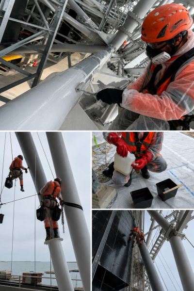 Workers in safety gear performing maintenance on large steel structures near the sea.
