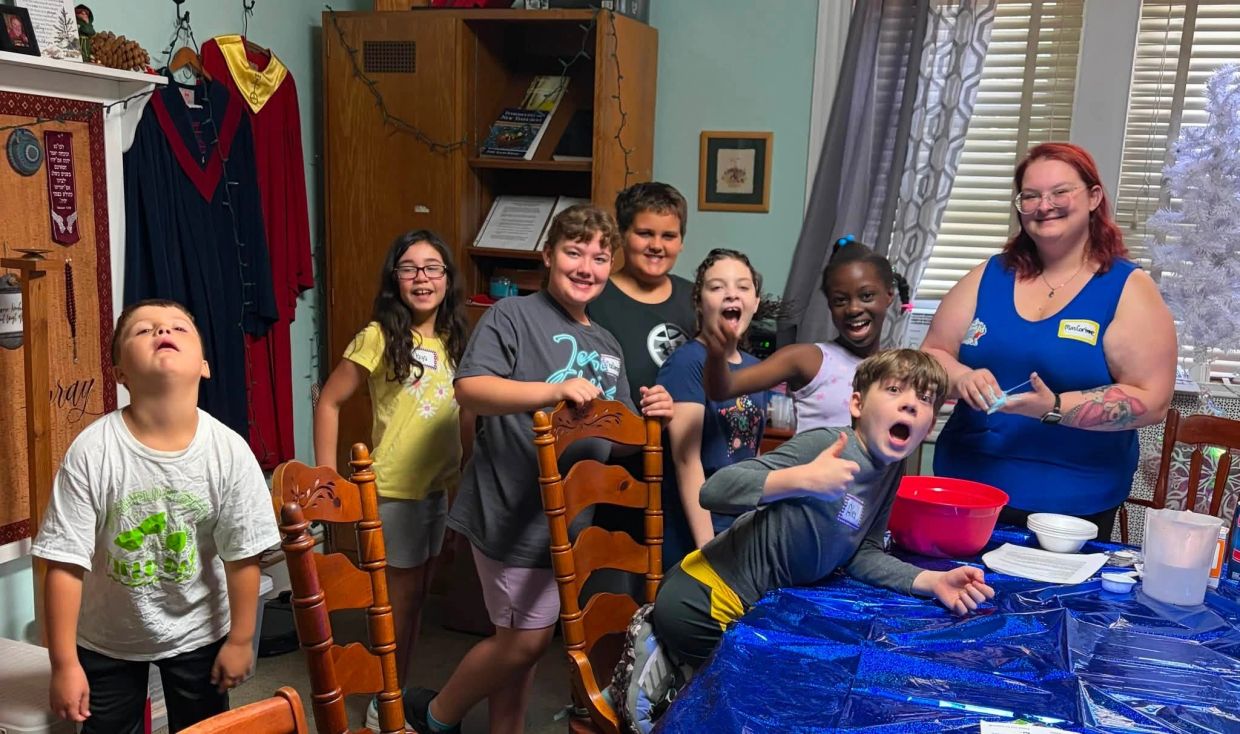 Group of children and a woman in a room around a table with a blue tablecloth.