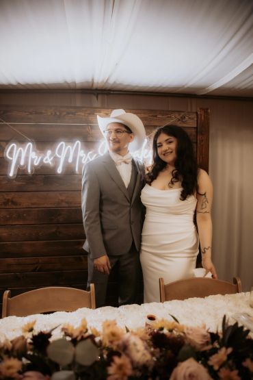 Newlywed couple posing by a neon "Mrs &amp; Mrs" sign at their wedding reception.