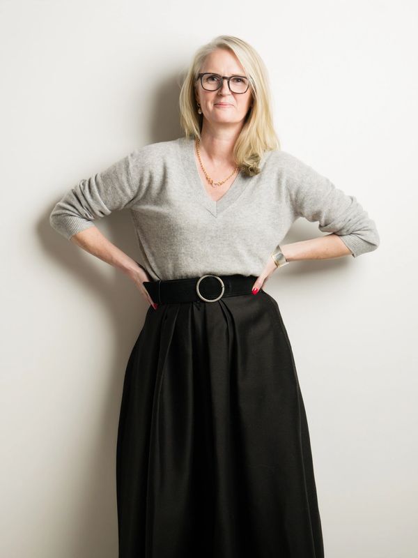 Confident middle-aged woman with glasses, wearing a gray sweater and black skirt, posing with hands on hips.