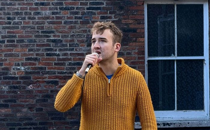 Man in mustard sweater speaking into a microphone outdoors.